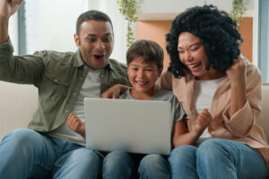 Happy family African American parents mum dad and Caucasian little boy kid child teenager son use laptop playing video game together watch fun cartoons win bet online victory celebrate on sofa at home