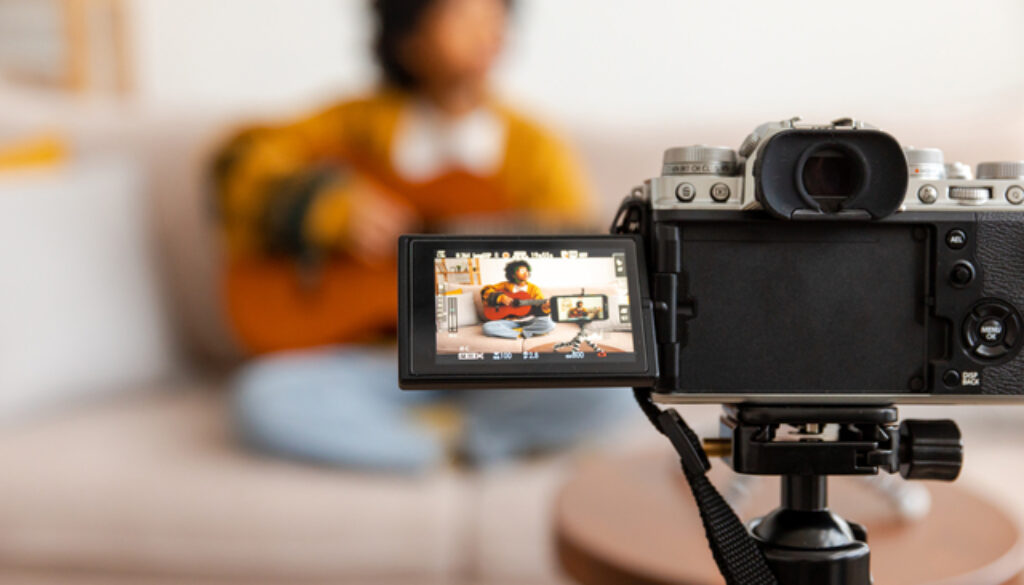 Blogger guitarist. African american girl blogger playing guitar talking to webcam recording vlog. Social media influencer woman streaming at home indoors. Music content creator broadcast tutorial.