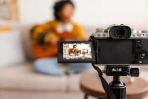 Blogger guitarist. African american girl blogger playing guitar talking to webcam recording vlog. Social media influencer woman streaming at home indoors. Music content creator broadcast tutorial.